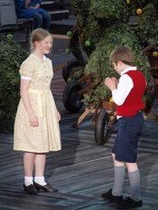 Young girl and boy in 1940s clothing. The boy is eating an apple as the girl grins and watches.